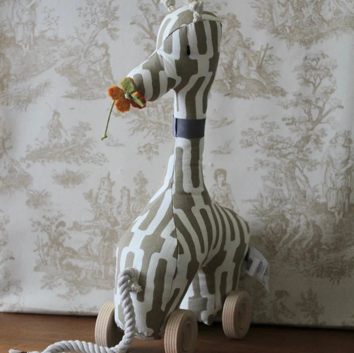 A giraffe heirloom pulltoy that is grey and white. It had a navy blue collae and a movable felt butterfly in it's mouth. Its on wooden weels and had a rope attached for pulling purposes. Baby Giraffe Pull-Toy, Wooden Toy for Toddlers, Nature-Inspired Wooden Pull Toy, Handcrafted Baby Giraffe Toy