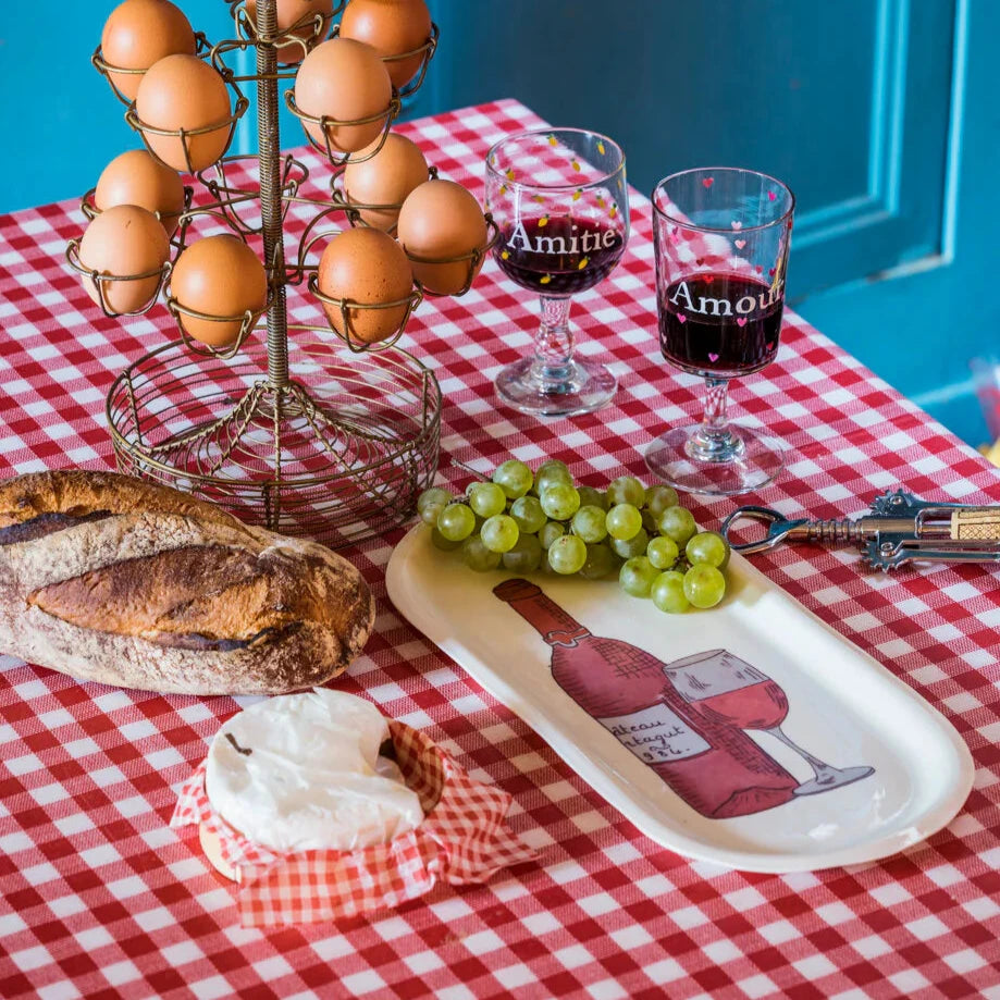 The Red Wine Bottle Tray features a hand-painted Wine glass and bottle illustration on high-quality enameled porcelain. This beautifully crafted tray, made using traditional plaster molds, showcases intricate craftsmanship and design. The tray is dishwasher safe for easy care and maintenance. The image highlights the elegant 14-inch length and 6-inch width of the tray, with the delicate illustration applied by hand in the Parisian workshops of Marin Montagut. This is seen in a realist place setting 